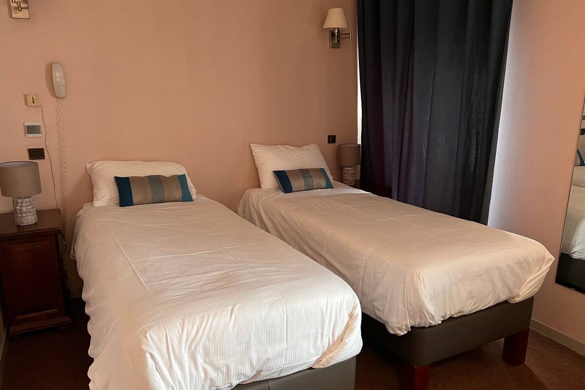Une chambre d’hôtel avec deux lits simples, une table de chevet avec une lampe, un téléphone fixé au mur et une fenêtre avec des rideaux.