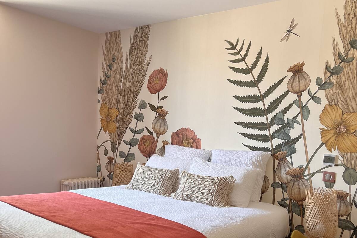 Une chambre à coucher avec un lit, une couverture rouge, une chaise jaune et une fresque murale représentant des fleurs et des feuilles.