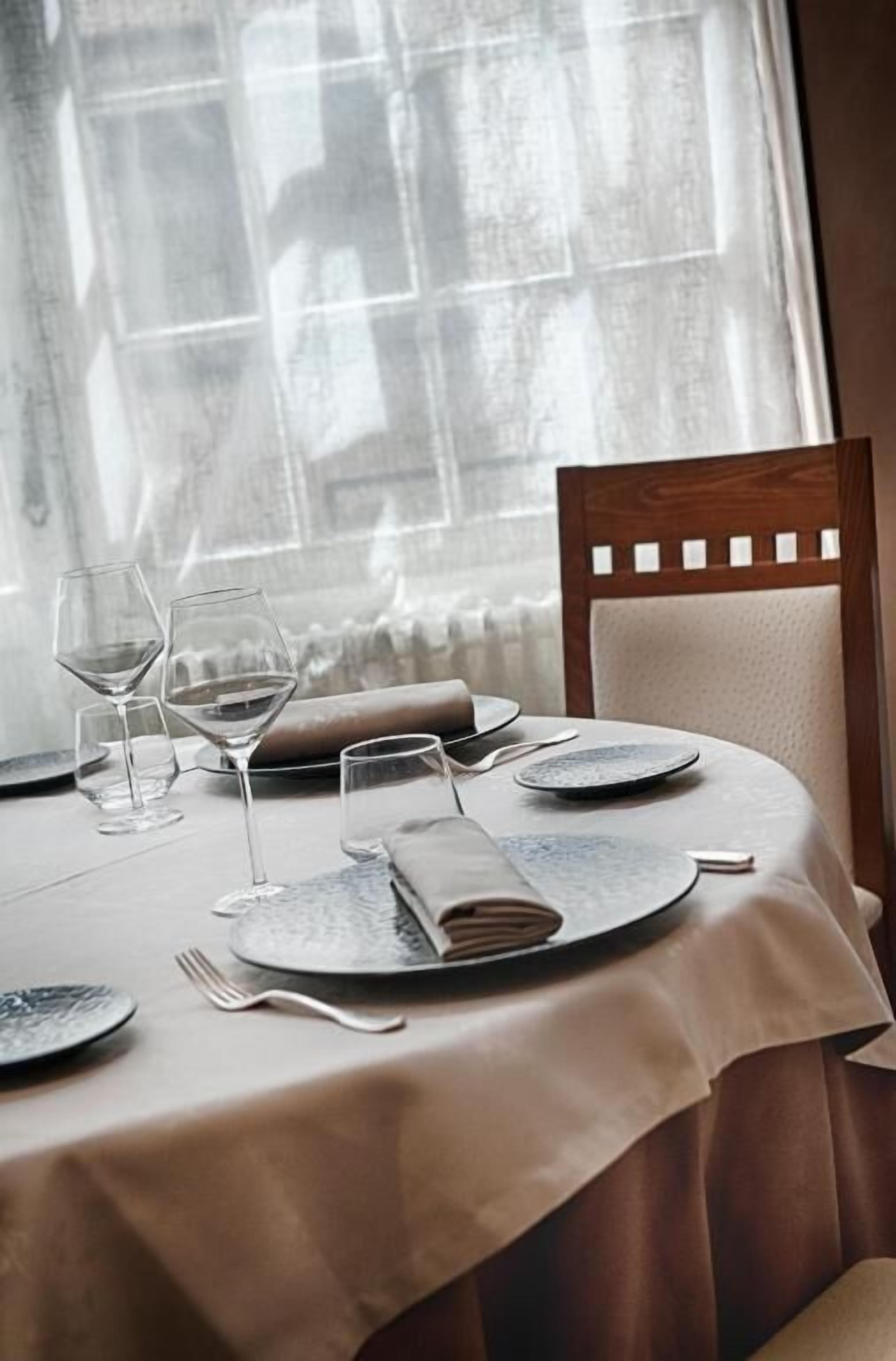 A dining table set with plates, glasses, and a book, with a chair and a window in the background.