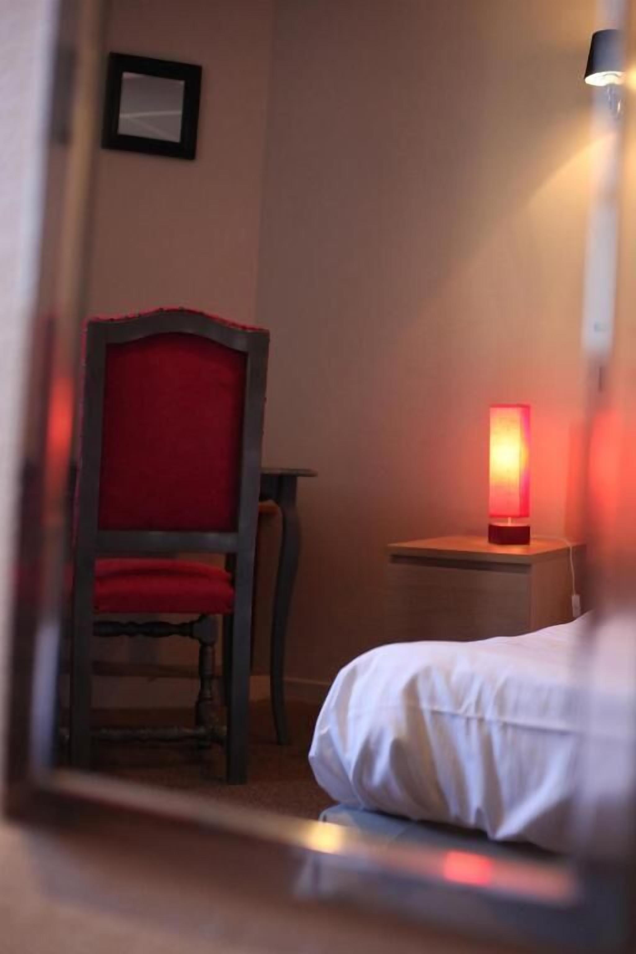 A hotel room with a bed, a chair, a nightstand with a red lamp, and a framed picture on the wall.