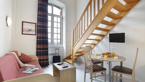 A modern room with a wooden staircase, a sofa, a small table with chairs, and a window with curtains. - Vacancéole - Les Demeures Champenoises (Epernay)