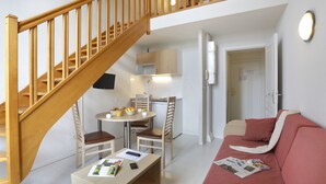 A compact living space with a wooden staircase, a dining area with a table and chairs, a kitchenette, a sofa, and a door leading to another room. - Vacancéole - Les Demeures Champenoises (Epernay)
