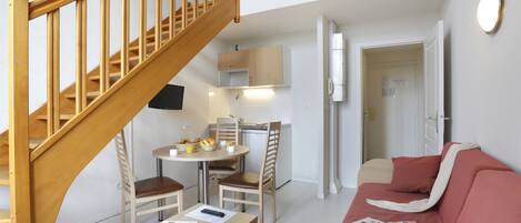 Een ruimtelijk ingerichte leefruimte met een houten trap, een eethoek met tafel en stoelen, een kitchenette, een bank en een deur naar een andere kamer.