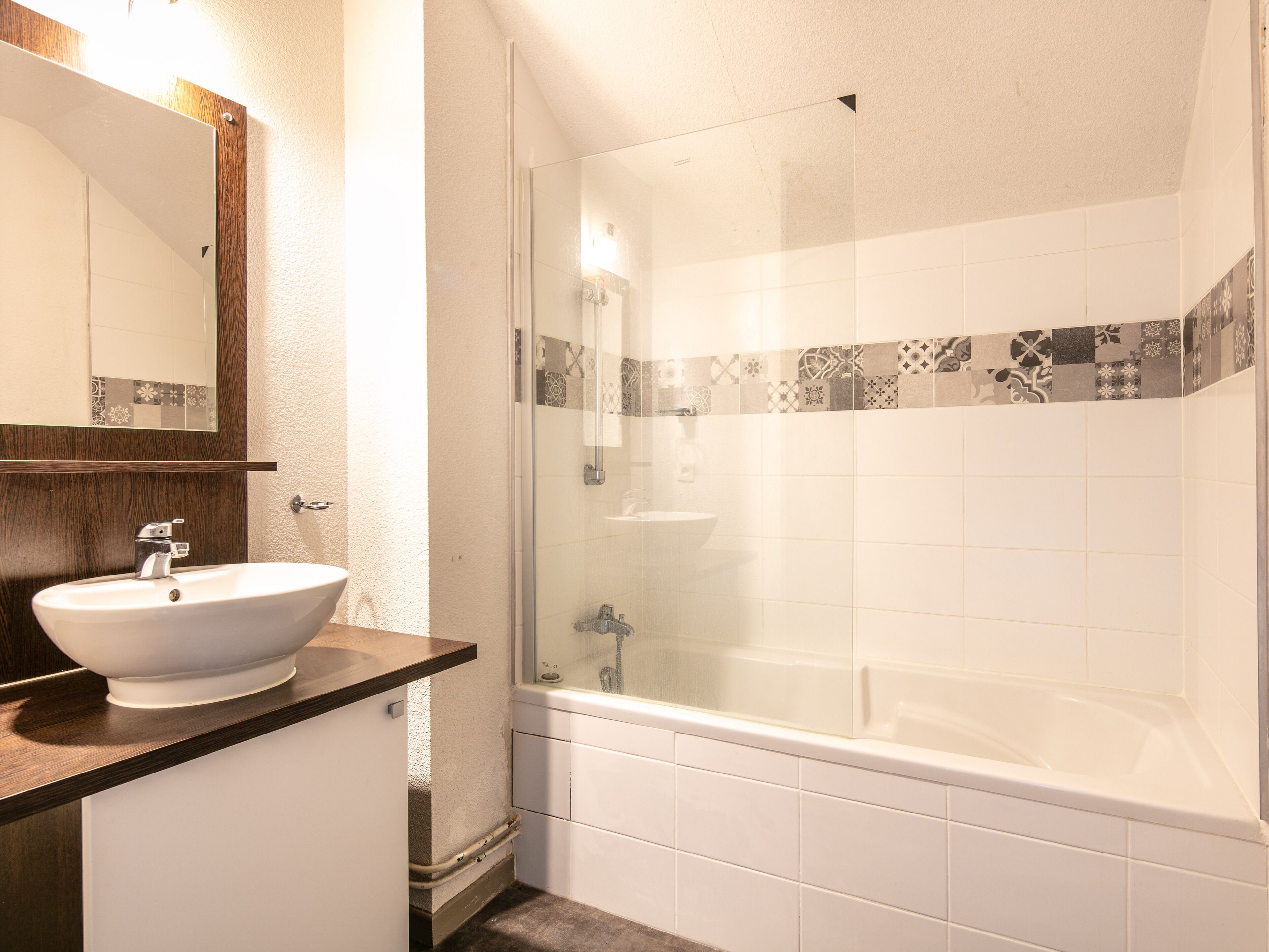 Une salle de bain avec un lavabo blanc, une douche à parois de verre et un mur recouvert de carreaux agrémenté d’une bordure décorative.