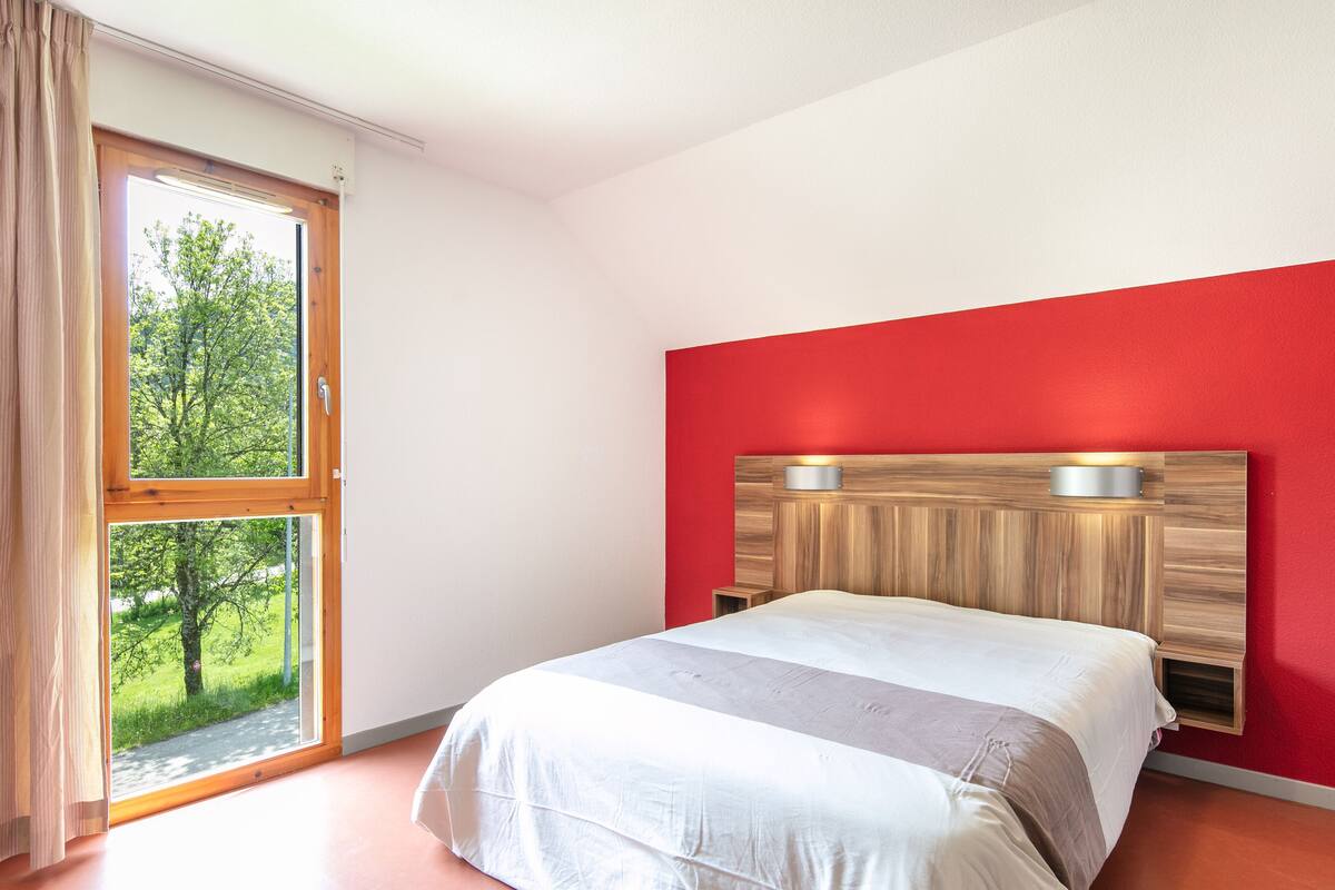 A bedroom with a large window, a red accent wall, and a wooden headboard.