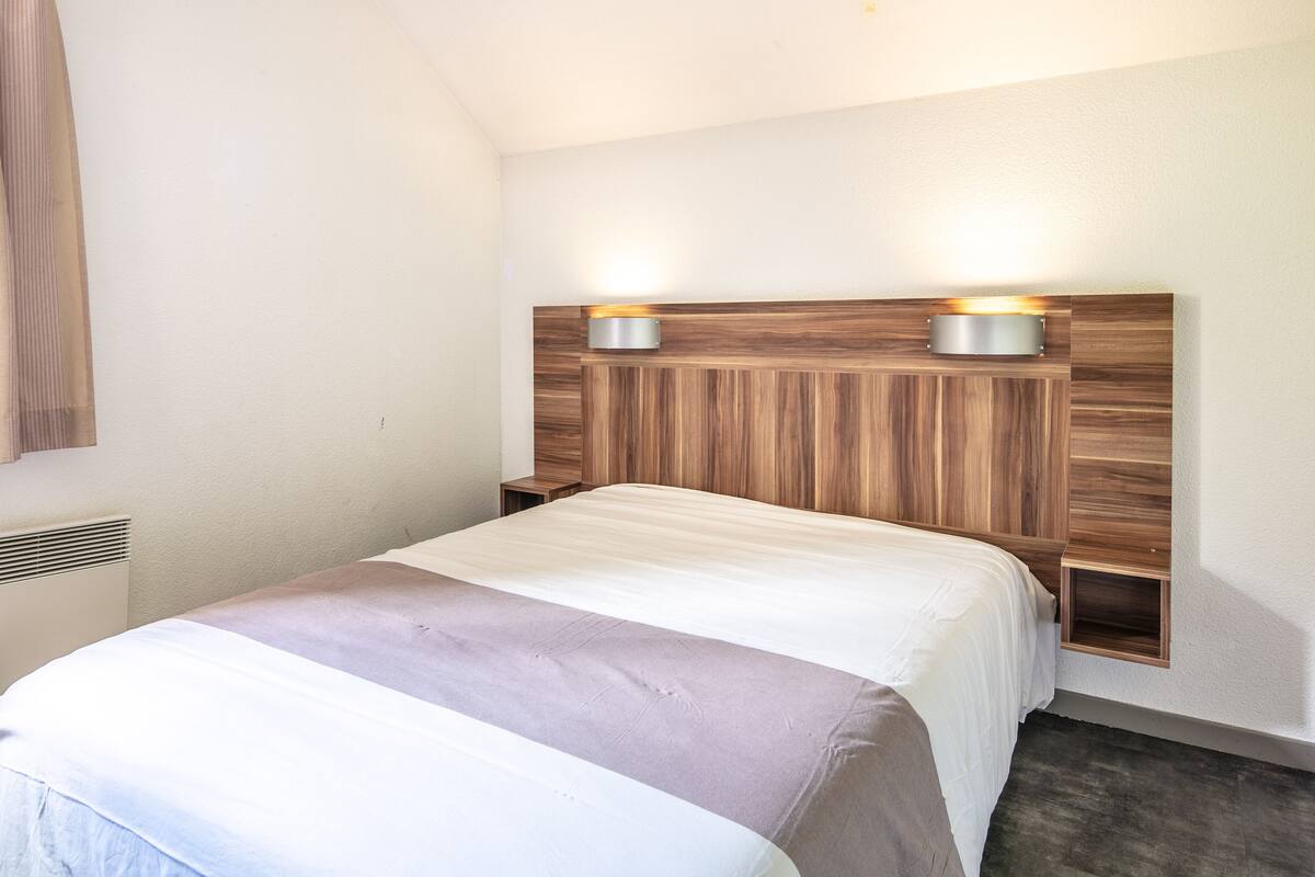 A hotel room with a wooden headboard, a bed with a white and beige bedspread, and two wall-mounted lights.