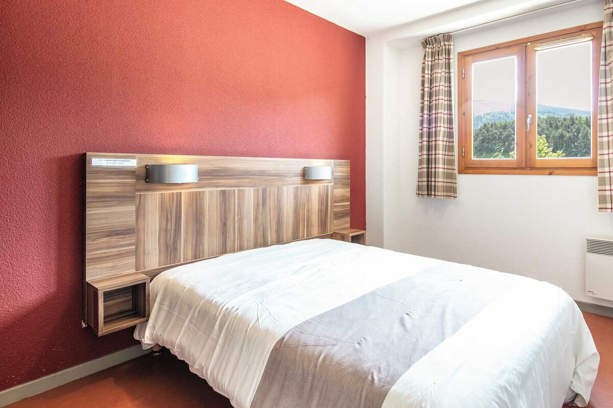 Une chambre avec une tête de lit en bois, un lit avec une literie blanche, une fenêtre avec des rideaux et un radiateur fixé au mur.