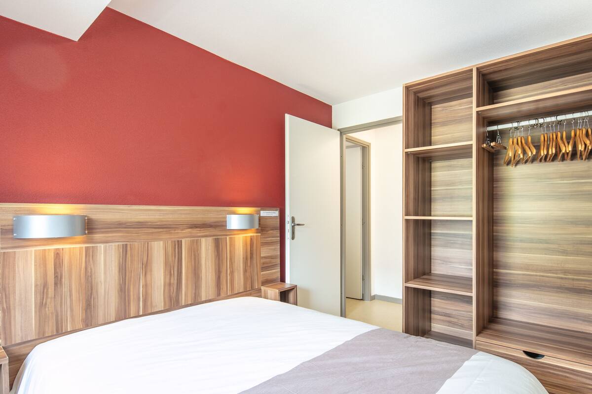 A modern hotel room with a large bed, a wooden headboard, a red accent wall, and a built-in wardrobe with hanging space.