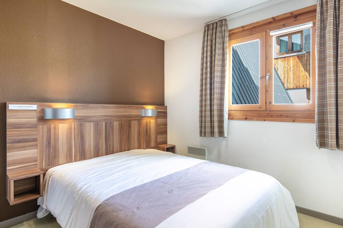A bedroom with a wooden headboard, a bed with white and grey bedding, a window with curtains, and a small shelf with a sign.