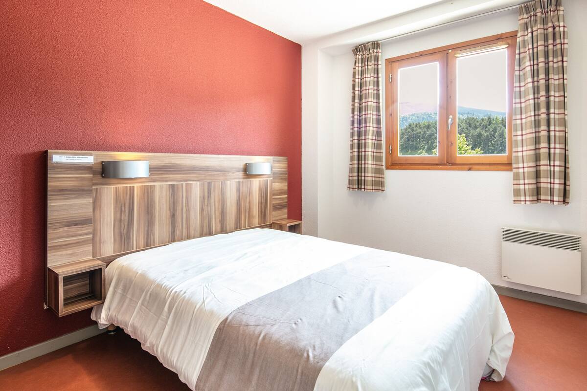 A bedroom with a wooden headboard, a bed with white bedding, a window with plaid curtains, and a radiator.