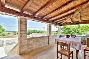 Une terrasse extérieure couverte, dotée de piliers en pierre, d’une table avec des chaises et offrant une vue sur la verdure et les bâtiments.