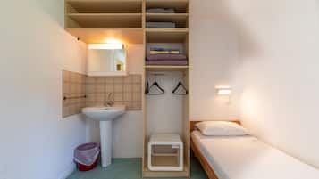 Une petite pièce avec un lavabo blanc, un miroir, un petit tabouret, un lit avec une literie blanche et une étagère où sont pliées des serviettes et des livres.