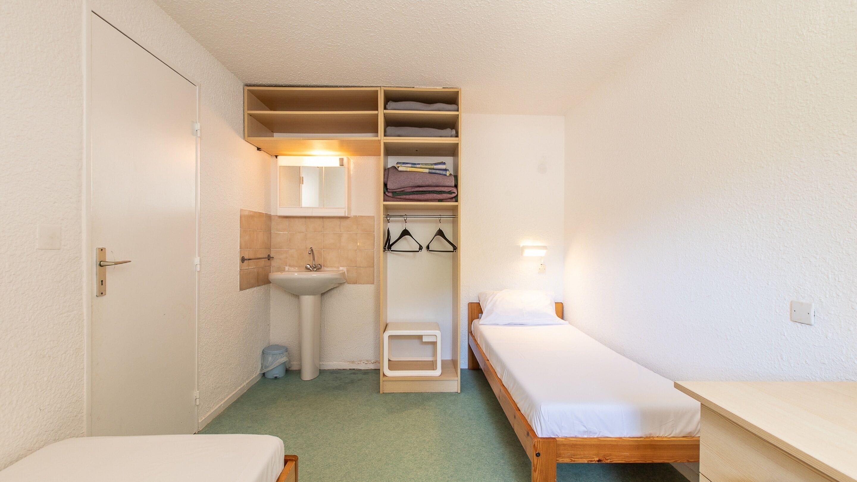 Una habitación pequeña y funcional con una cama individual, una mesita de noche, armario empotrado con estantes y un lavamanos.