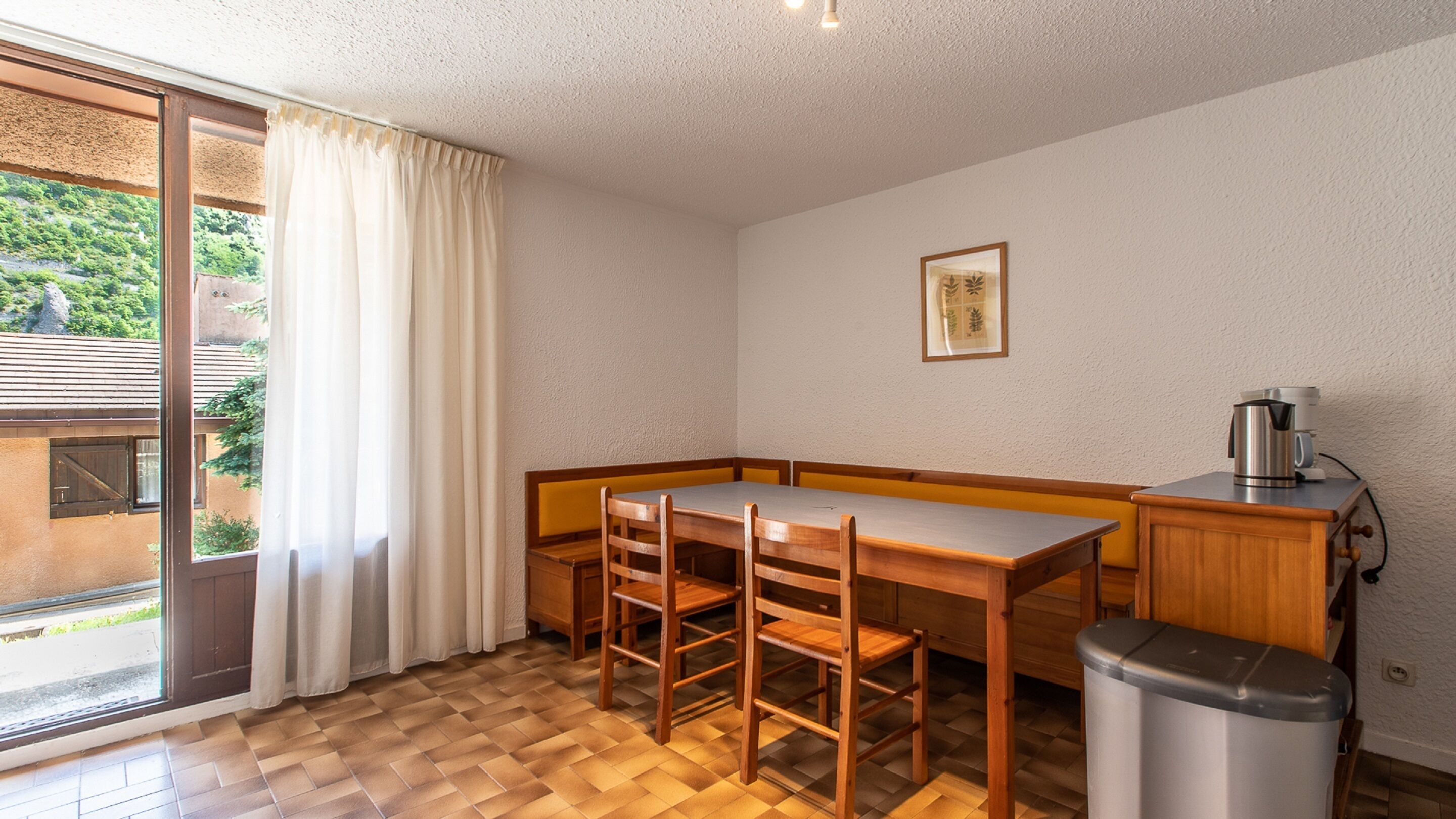 A small dining area with a wooden table and chairs, a trash bin, and a coffee maker.