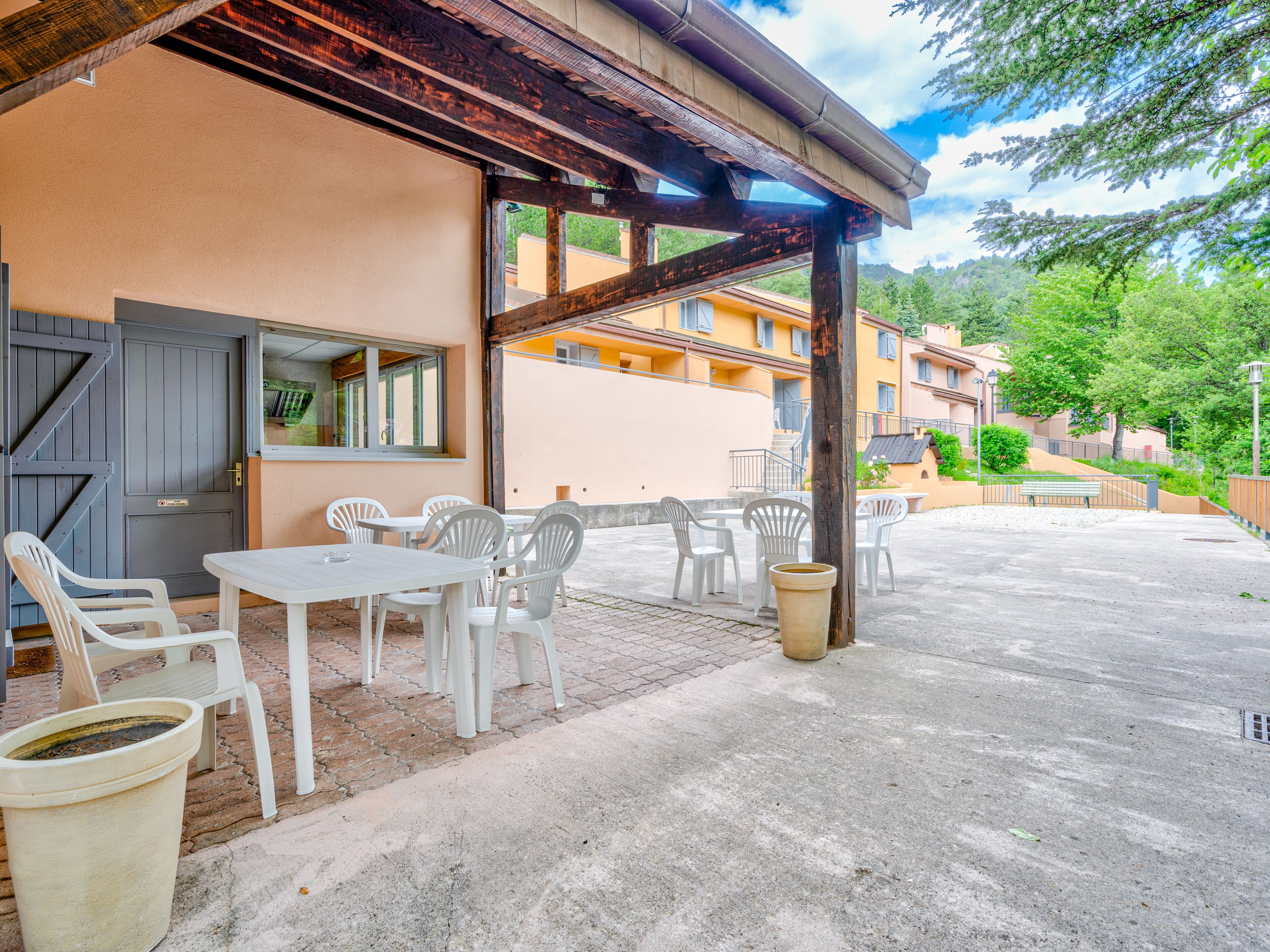 An outdoor seating area with white plastic chairs and tables, a concrete floor, and a building with a wooden pergola.