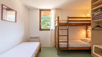 Een kleine, gezellige kamer met een stapelbed, een eenpersoonsbed, een raam met gordijnen, een radiator en een spiegel aan de muur.