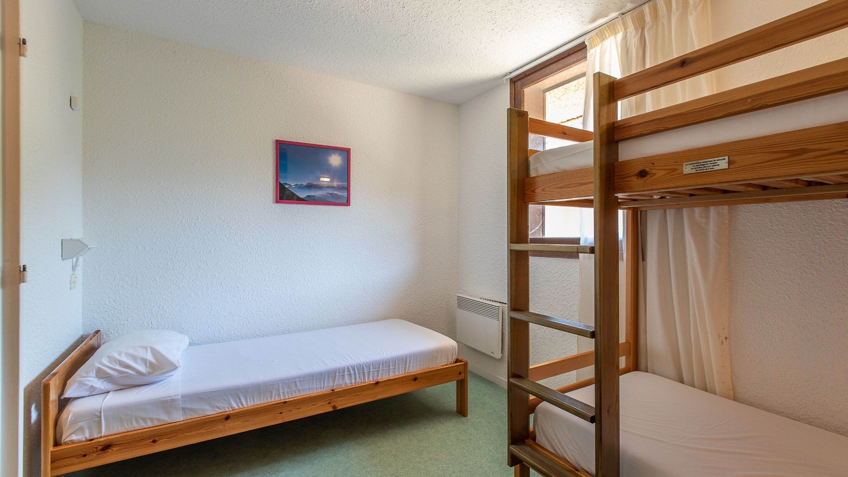 Une chambre avec un lit simple et un lit superposé, un tableau encadré au mur et une fenêtre avec des rideaux.
