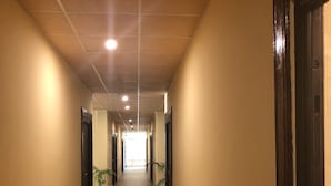 Un pasillo largo y estrecho con paredes beige y piso de baldosas. Hay puertas a ambos lados del pasillo, y macetas con plantas están colocadas a intervalos regulares. El techo tiene iluminación empotrada.