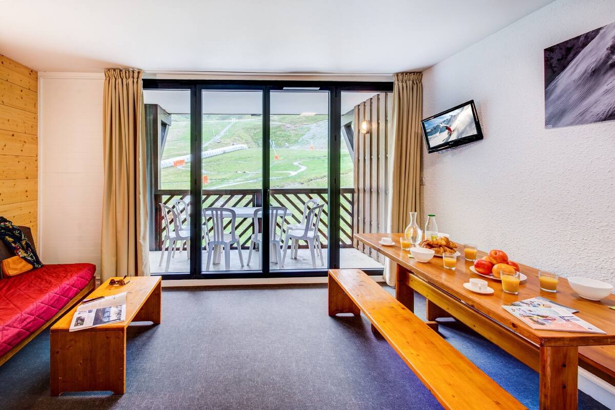A room with a red sofa, wooden benches, a breakfast table with fruit and drinks, and a large window with a view of a green landscape.
