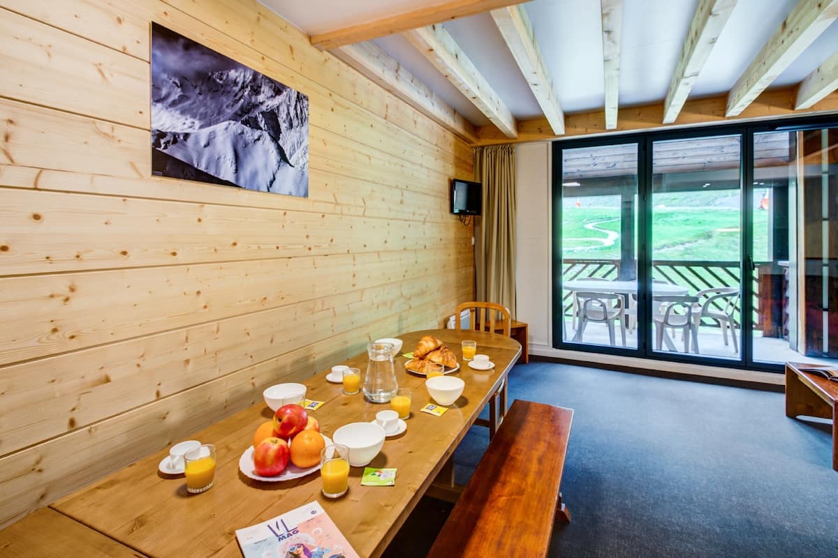 A wooden-paneled room with a breakfast table set with fruits, juice, and croissants. A television is mounted on the wall, and there is a large window offering a view of the outdoors.
