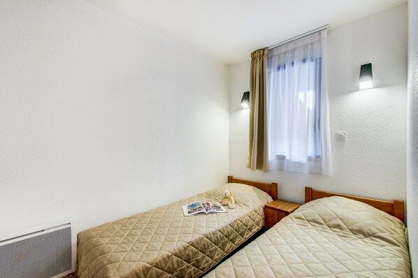 Una habitación pequeña y limpia con dos camas individuales, una ventana con cortinas y una lámpara fijada a la pared.