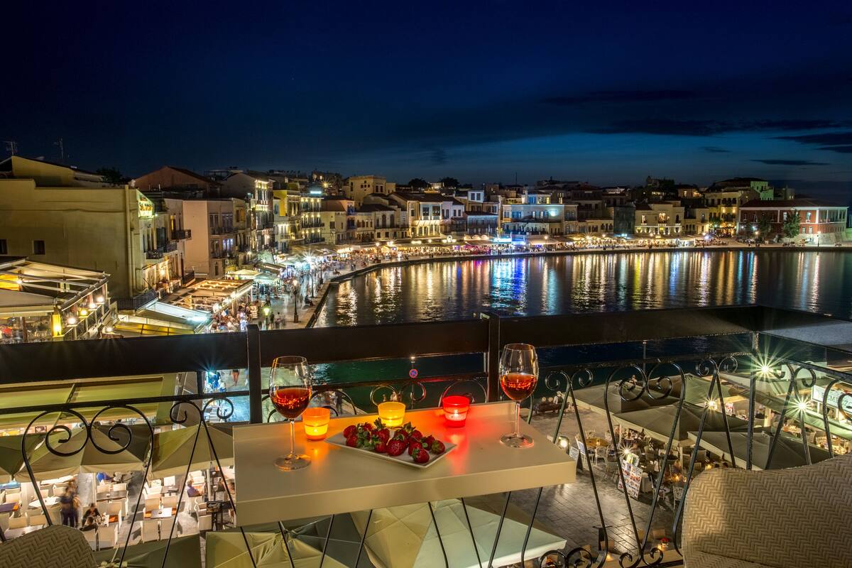 A terrace with a table set for two, overlooking a waterfront with buildings and a reflective water surface.