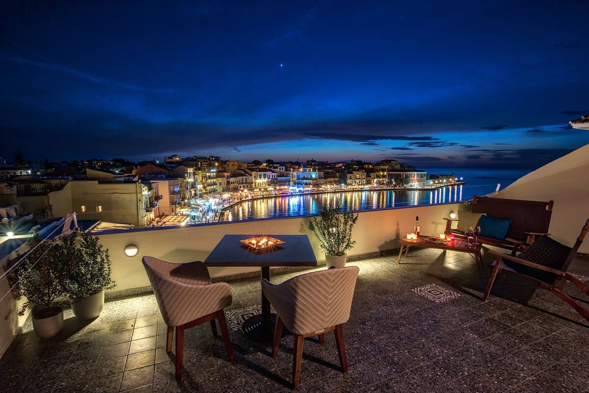 A rooftop terrace with a table and chairs, overlooking a waterfront with buildings and a pier.