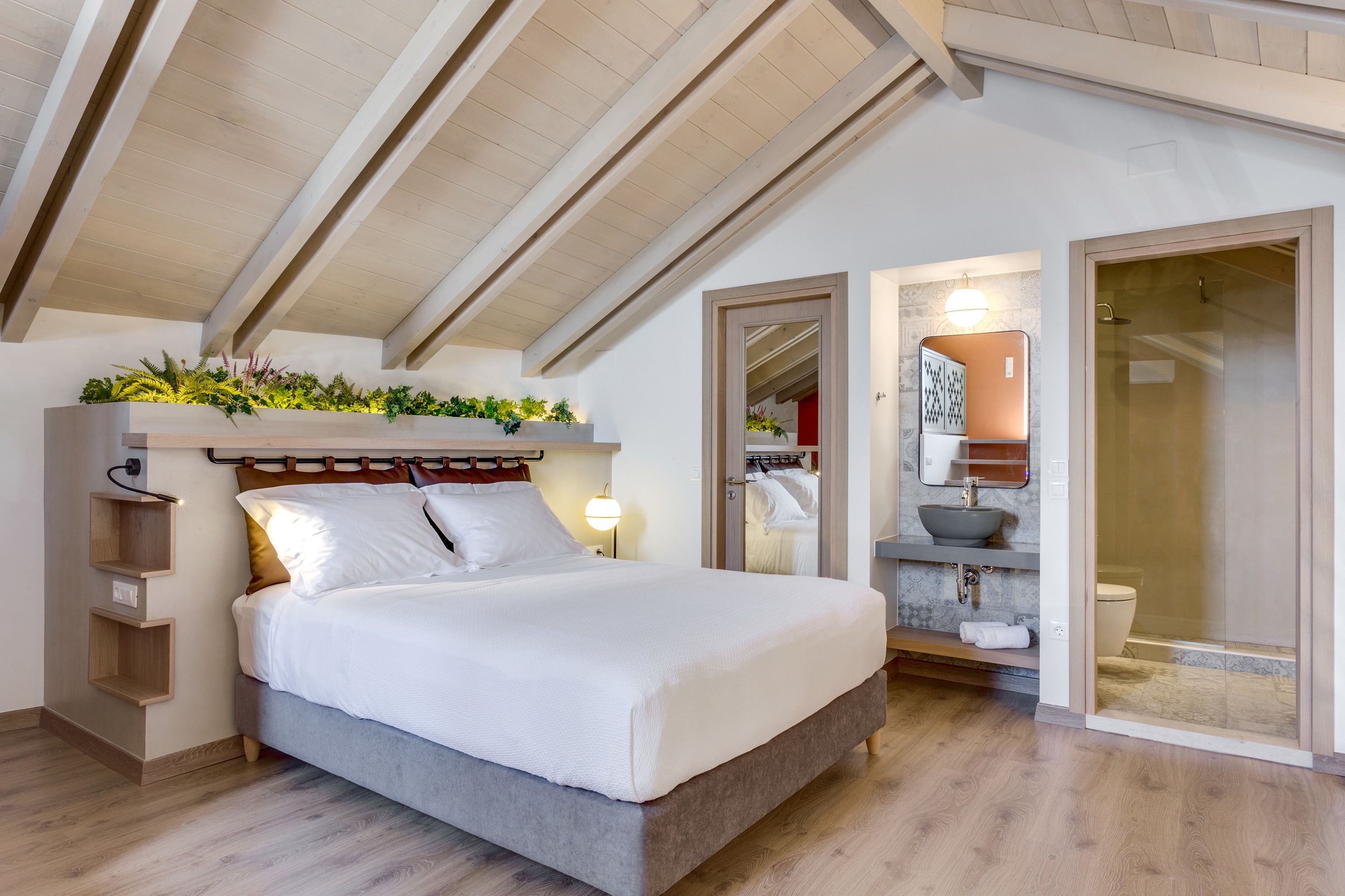 A modern bedroom with a large bed, wooden beams on the ceiling, a bedside plant arrangement, a built-in shelf, and a bathroom with a glass shower.