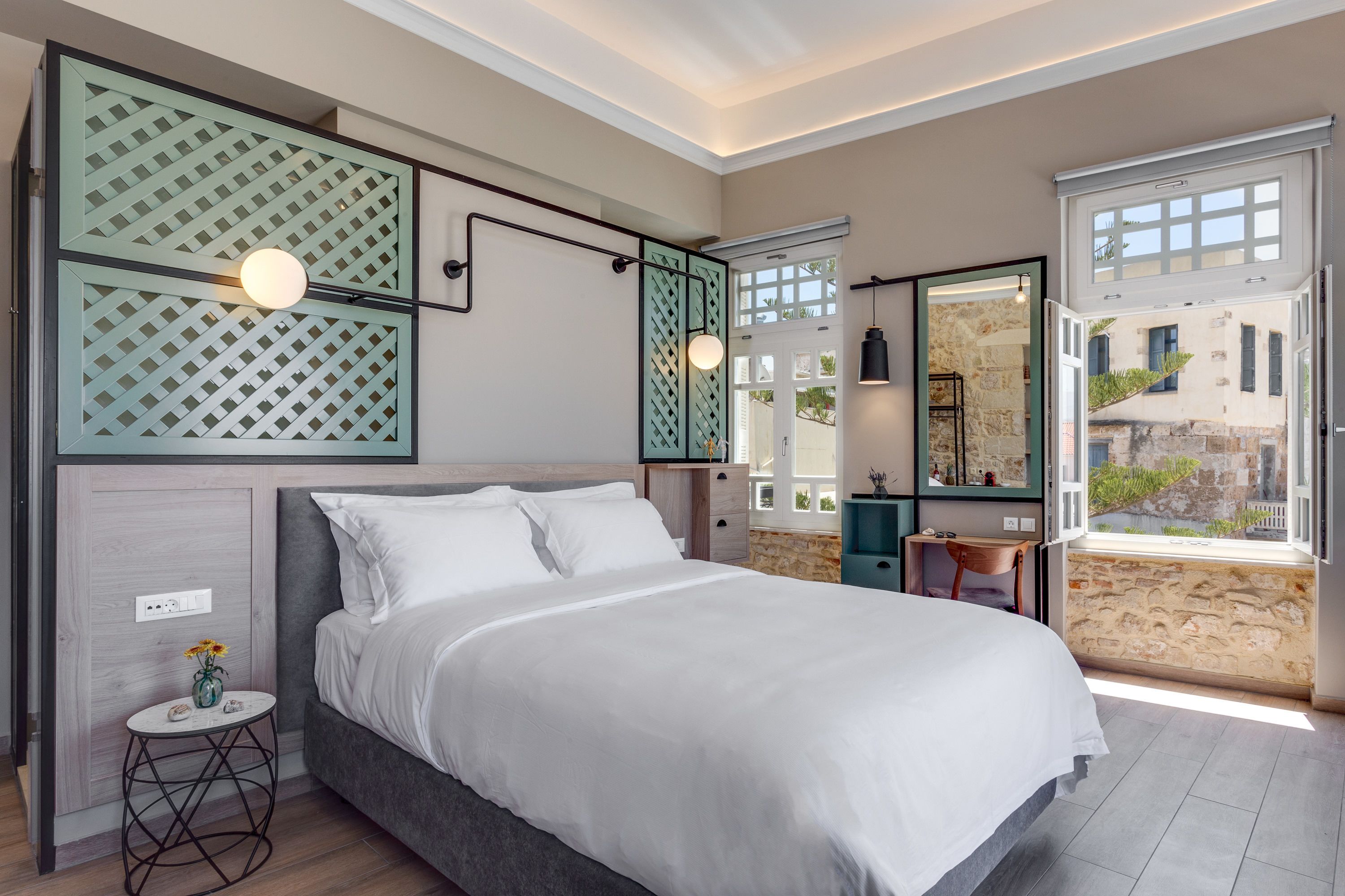A modern bedroom with a large bed, a small side table, a mirror, and a view of the outside.