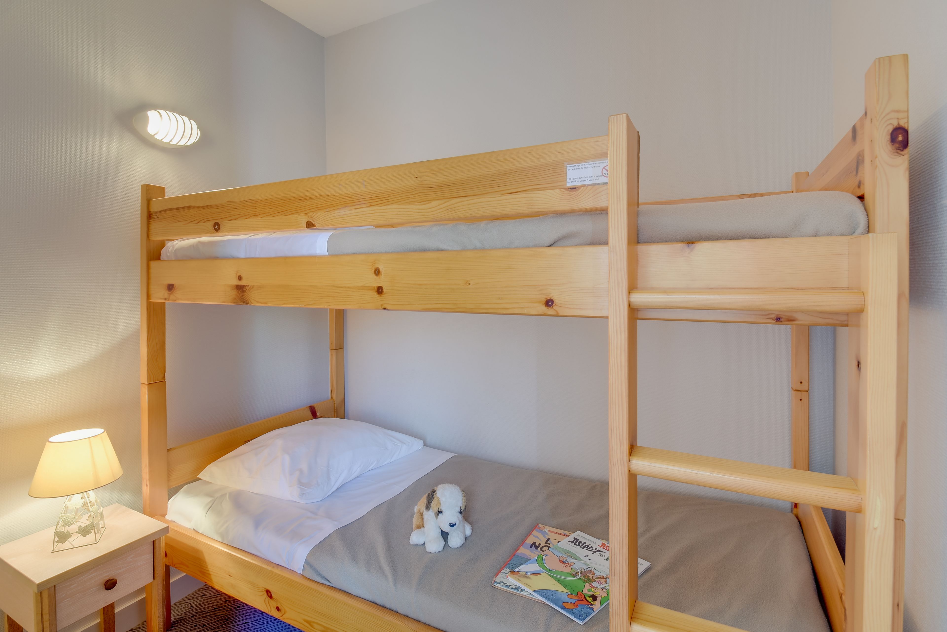 A bunk bed with a wooden frame, a nightstand with a lamp, and a plush toy on the lower bunk.