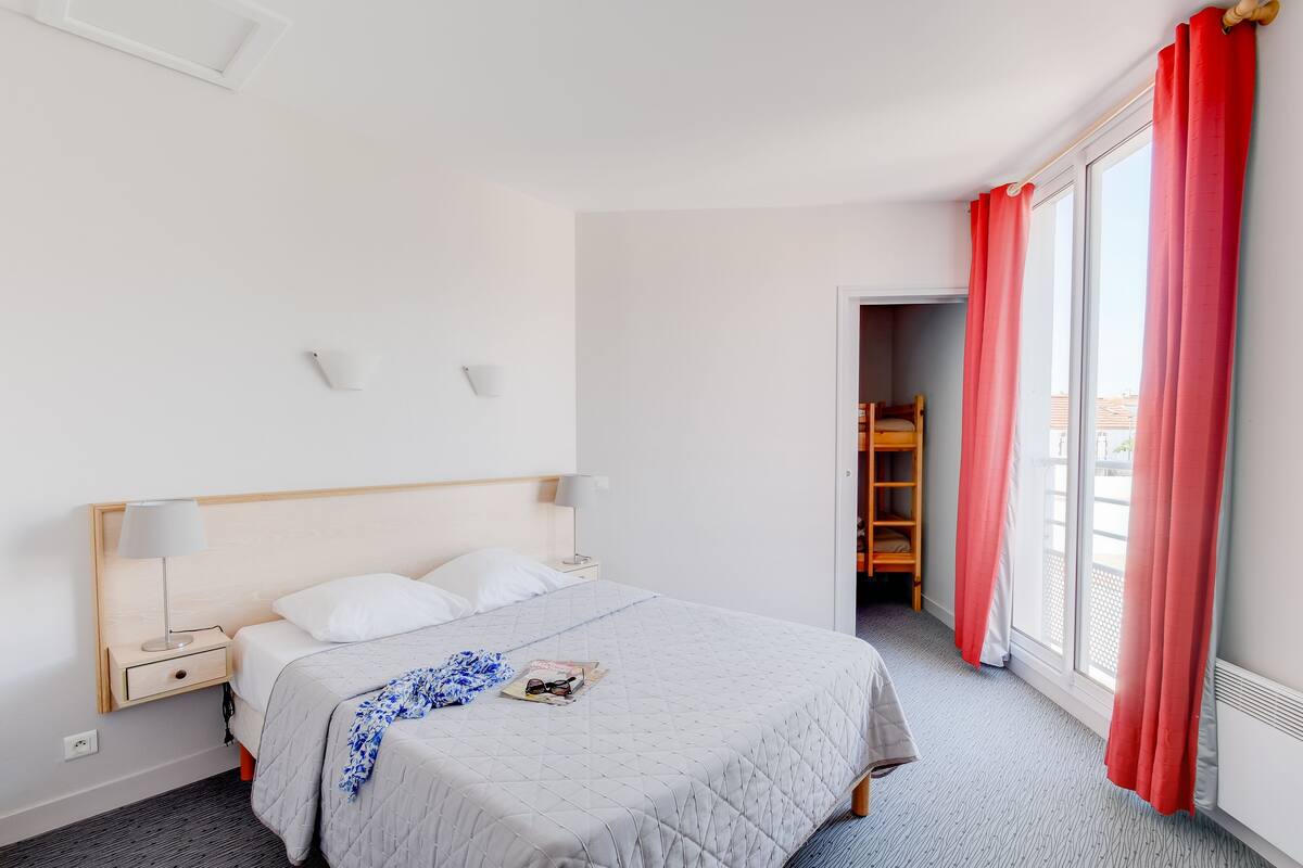 A hotel room with a bed, bedside tables, a lamp, a magazine, and a window with red curtains.