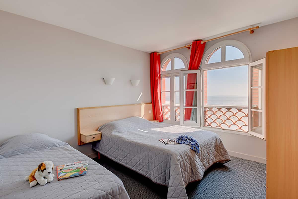 A hotel room with two beds, a window with red curtains, and a view of the sea.