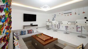 Ein modernes Wohnzimmer mit einer Couch, einem Couchtisch und einem an der Wand befestigten Fernseher. Drei weiße Sessel und ein kleiner Tisch mit einem dekorativen Gegenstand sind ebenfalls vorhanden.