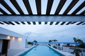 Zona de piscina con vista al mar, pérgola de madera y edificio blanco.