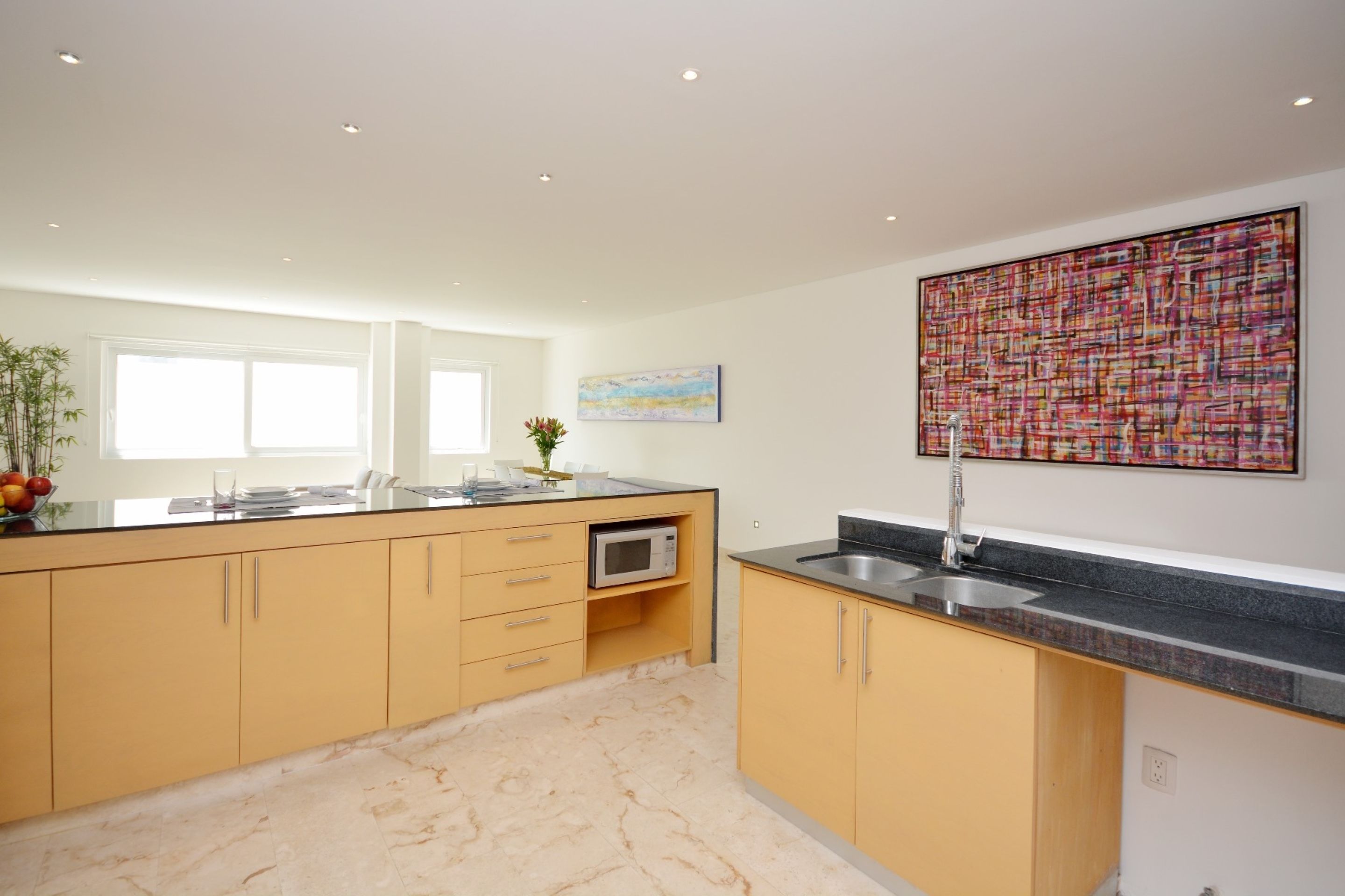 A modern kitchen with yellow cabinets, a black countertop, and a stainless steel sink. There is a microwave on the counter and a painting on the wall.