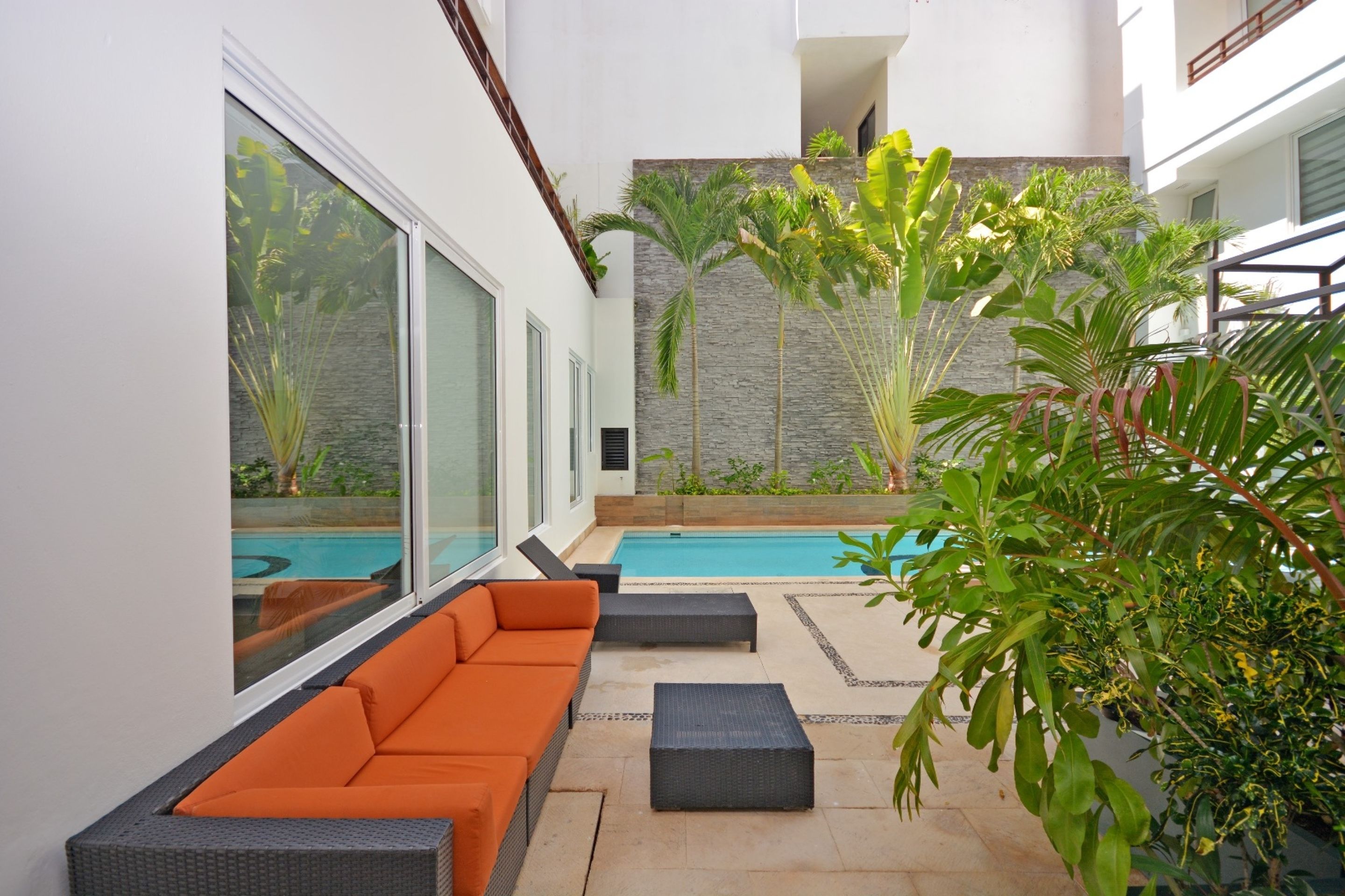 A modern outdoor patio with a pool, orange seating, and tropical plants.