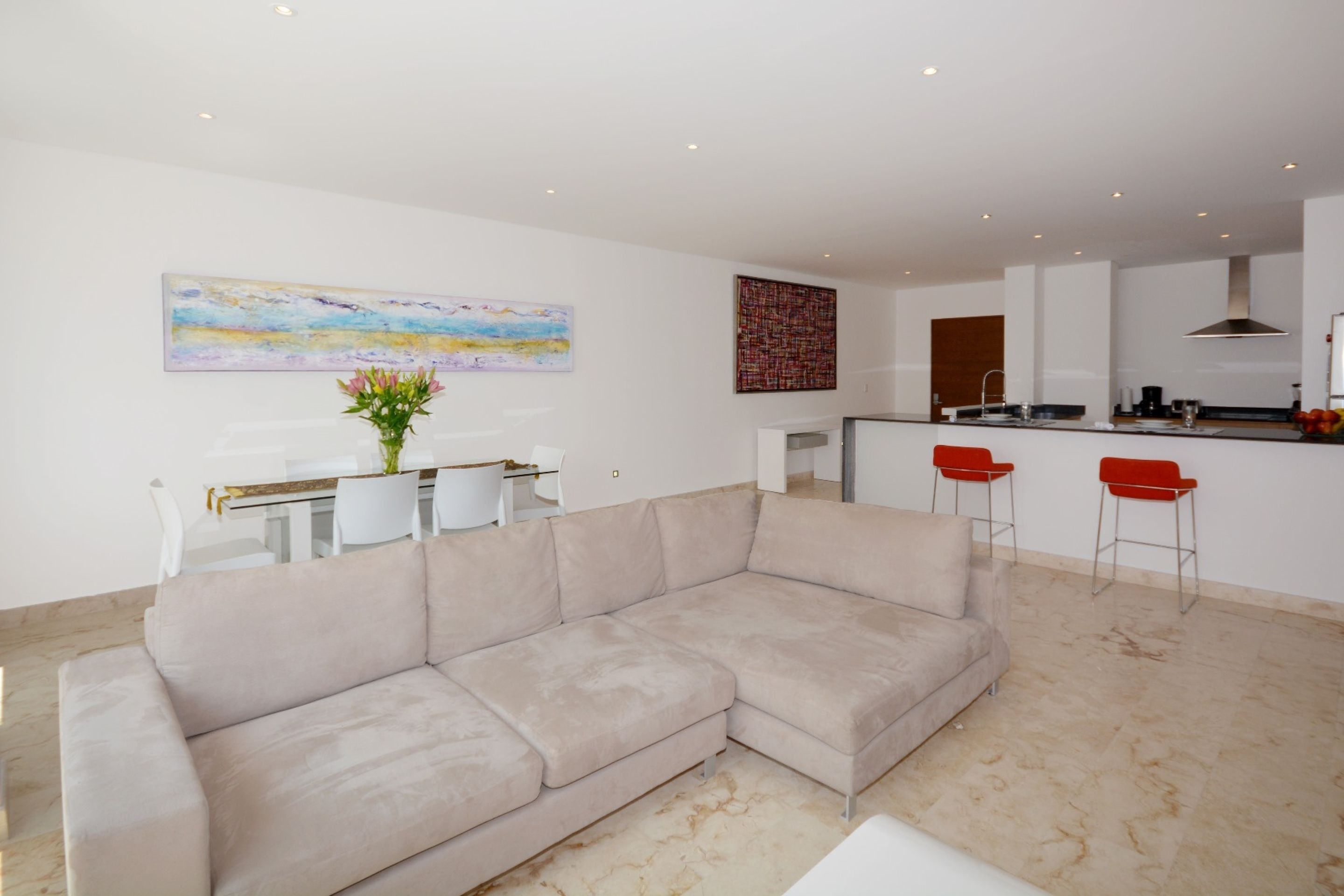 A modern living room with a beige sectional sofa, a dining area with a white table and chairs, and a kitchen area with a bar counter and red bar stools.