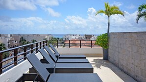A rooftop terrace with lounge chairs, a palm tree, and a view of the sea and buildings. - Stylish 2BR a few steps to the beach with pools by Happy Address (Playa del Carmen)