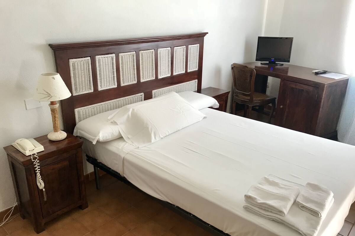 Una cama bien hecha con ropa de cama blanca, una mesita de noche de madera con una lámpara y un teléfono, un televisor sobre un escritorio y un cabecero de mimbre.