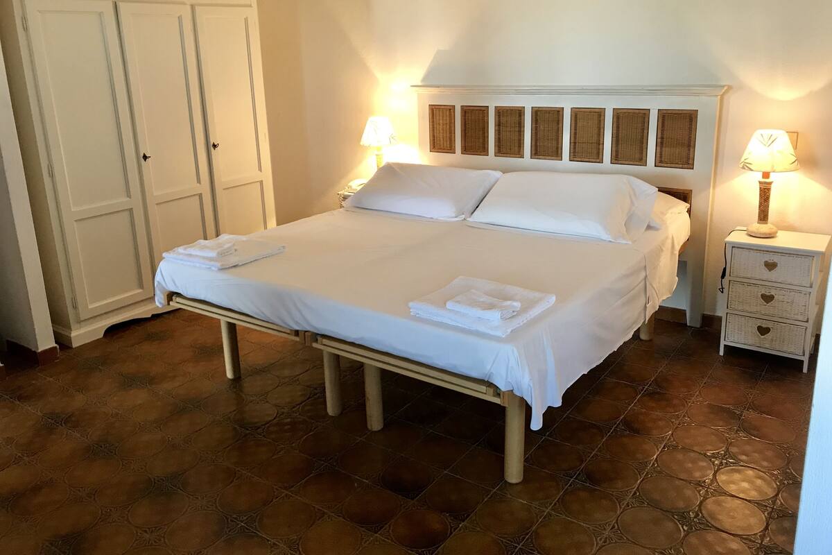 Una cama bien tendida con sábanas blancas y toallas dobladas, una mesita de noche con lámpara y un armario.