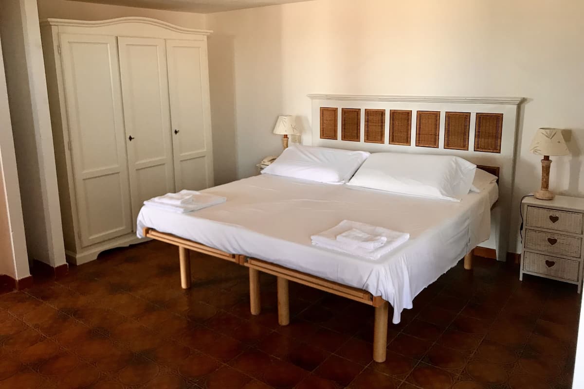 Una cama doble bien hecha con sábanas blancas, una mesita de noche con una lámpara y un armario con un tirador con forma de corazón.