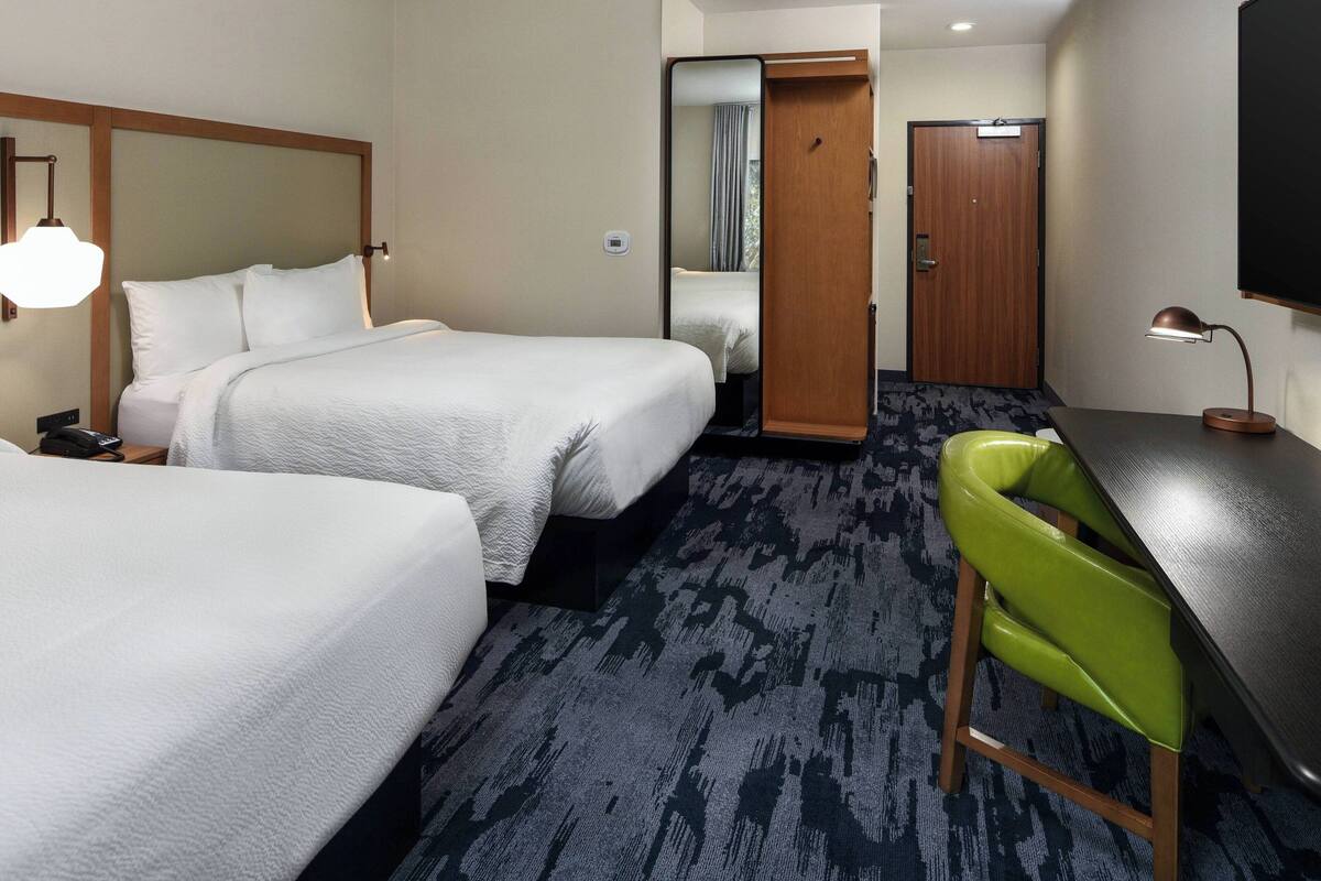 A hotel room with two beds, a desk, a green chair, a mirror, and a door.