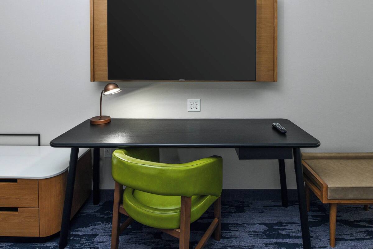 A room with a desk, a green chair, a small table, and a television mounted on the wall.