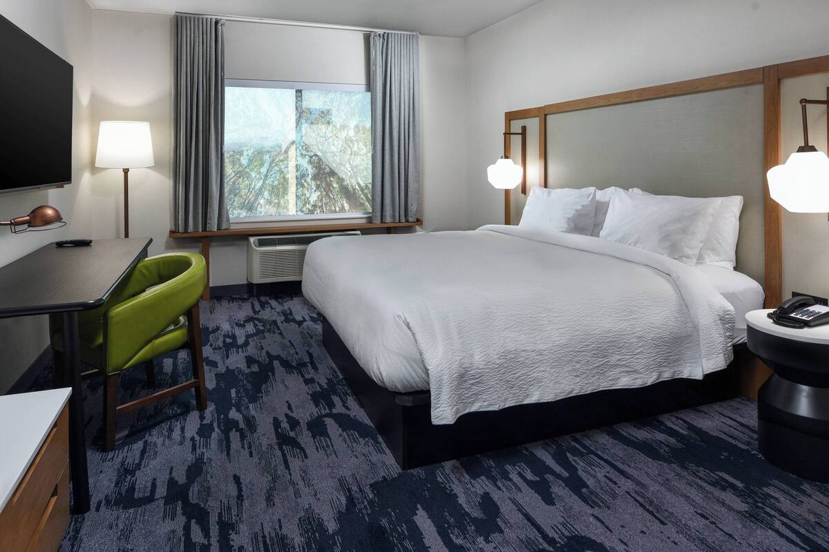 A hotel room with a large bed, a desk with a chair, a TV, and a window with curtains.