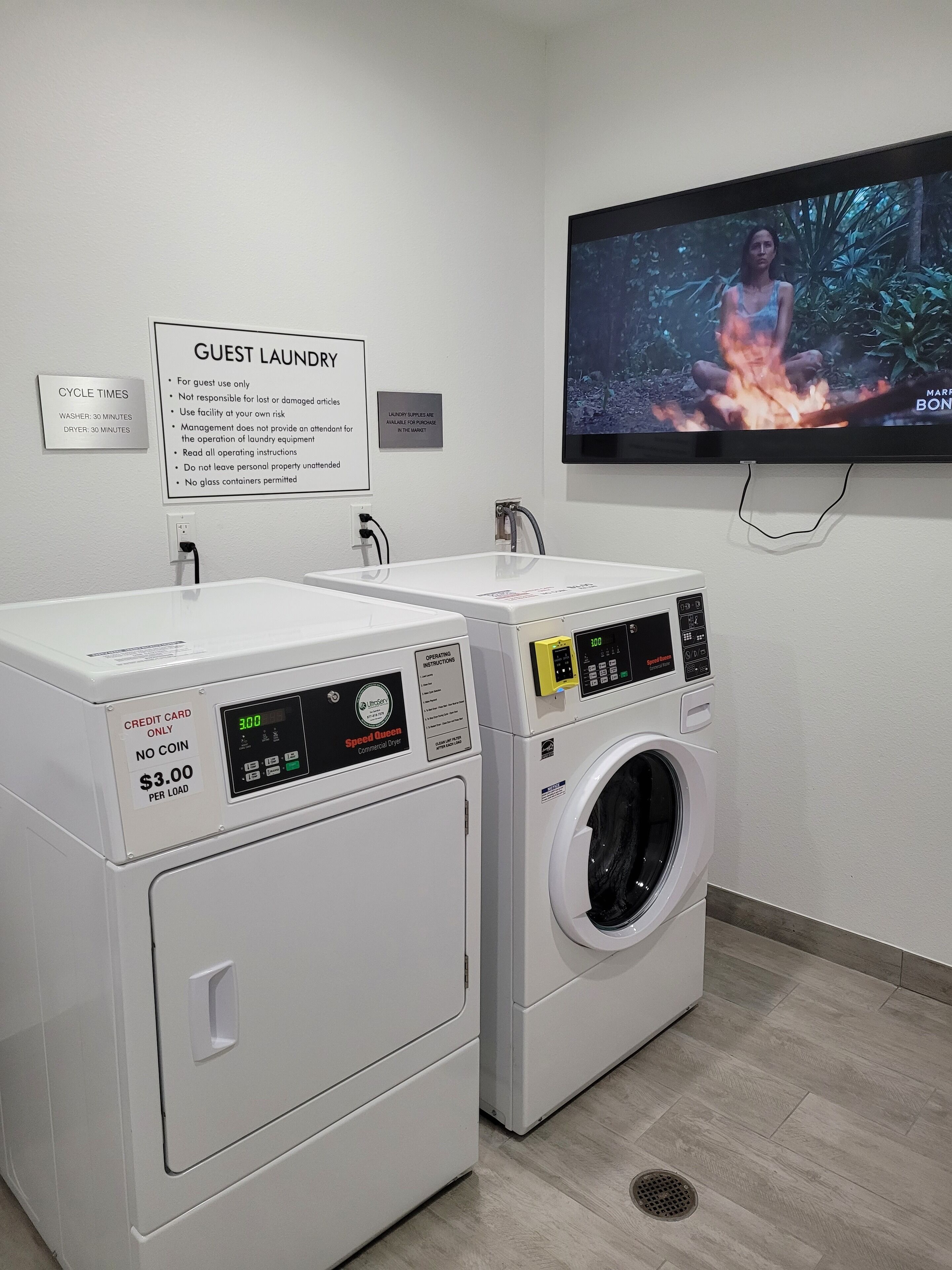 Zona de lavandería para huéspedes con dos lavadoras y una secadora, un cartel con instrucciones de uso y un televisor que muestra una escena de naturaleza.