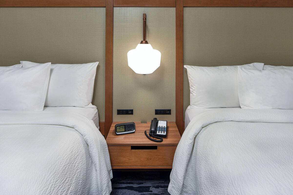 A hotel room with two beds, a bedside table with a lamp, a phone, and a digital clock.