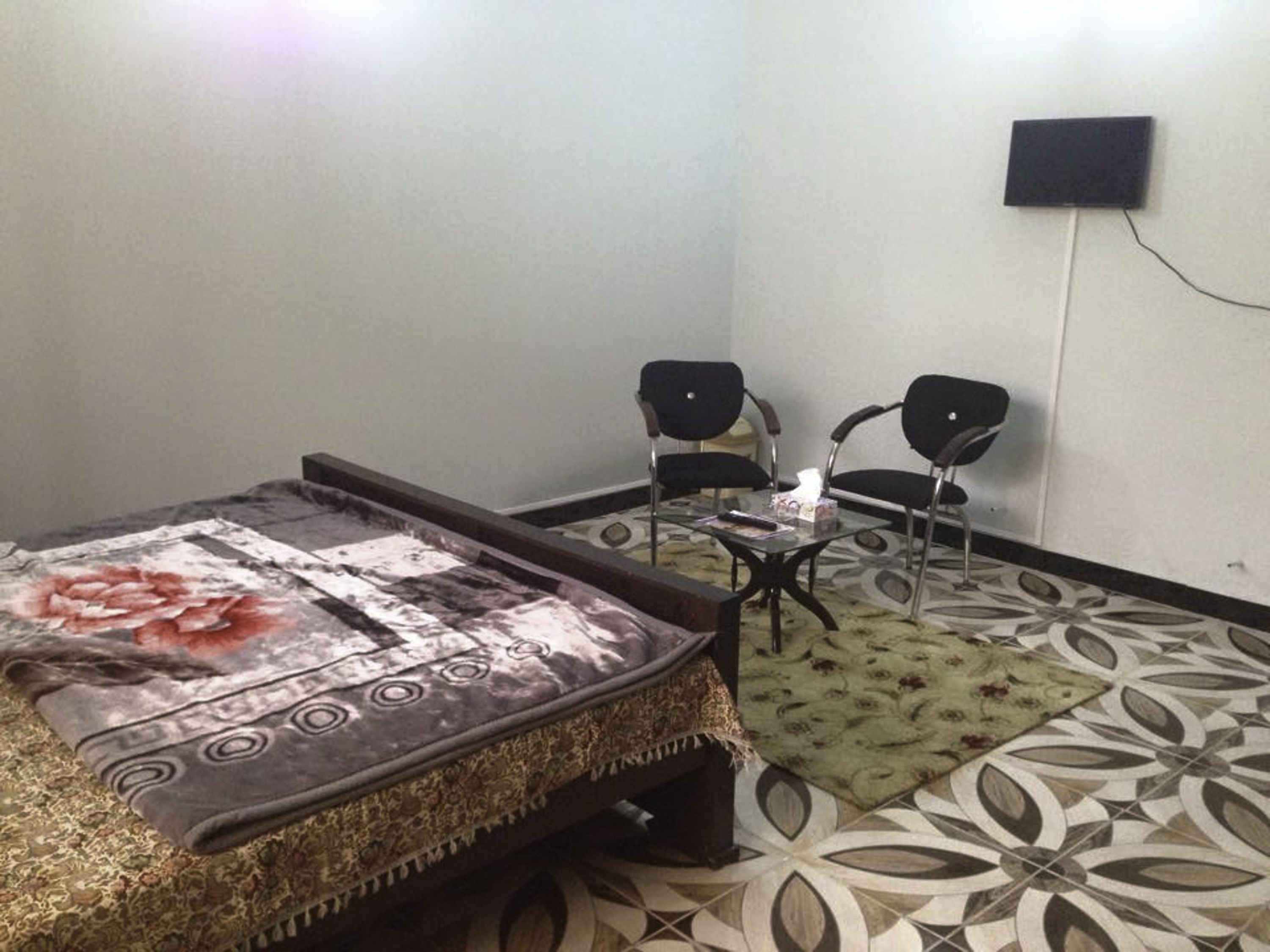 Um quarto pequeno com uma cama, duas cadeiras, uma mesa e uma TV fixada na parede.