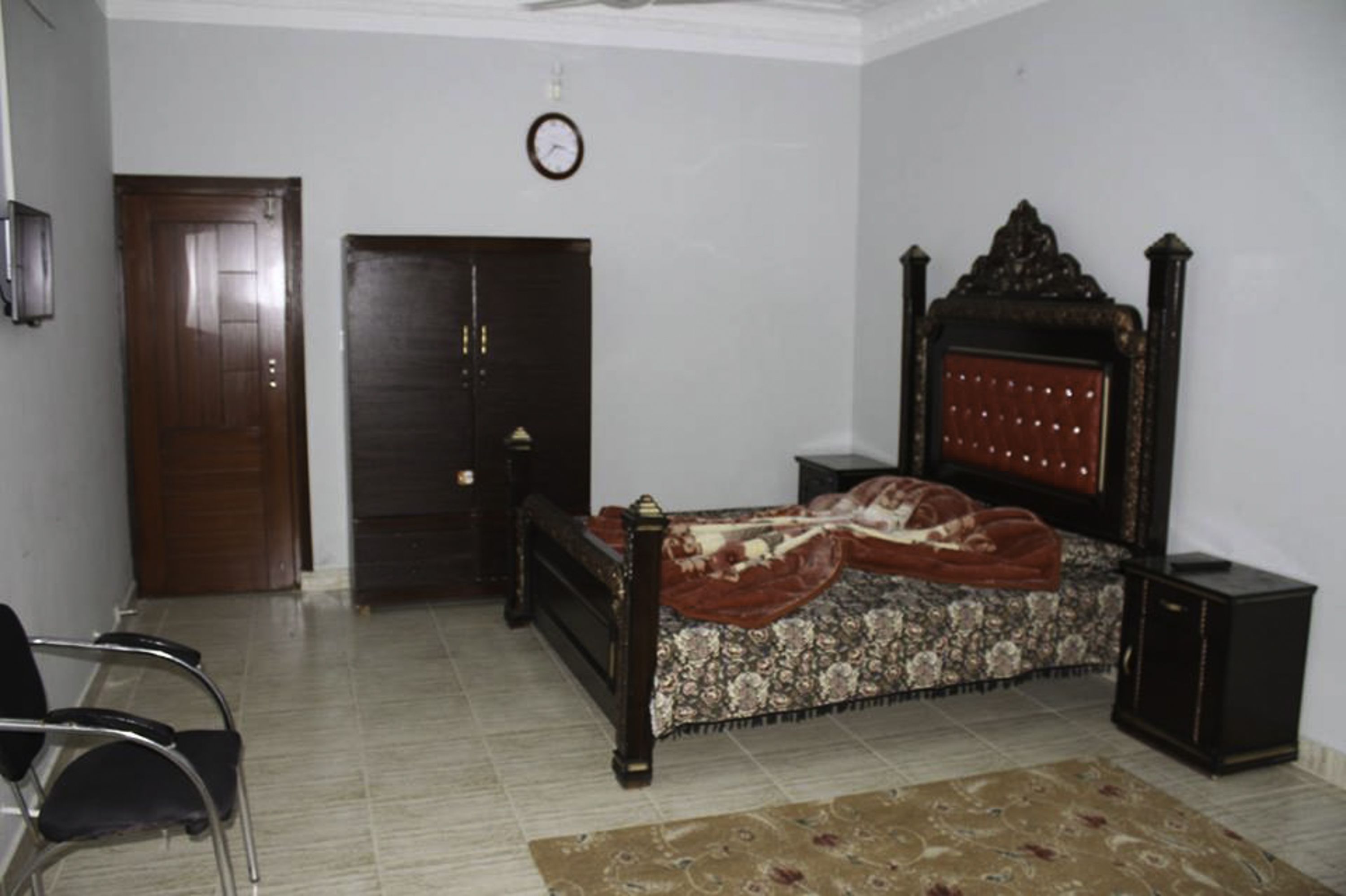 Una camera da letto con un letto, una porta in legno, un orologio appeso al muro e un comodino accanto al letto.