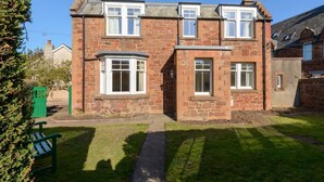 A two-story stone house with multiple windows, a paved driveway, and a green bench. - Glenconner Garden Cottage (North Berwick)