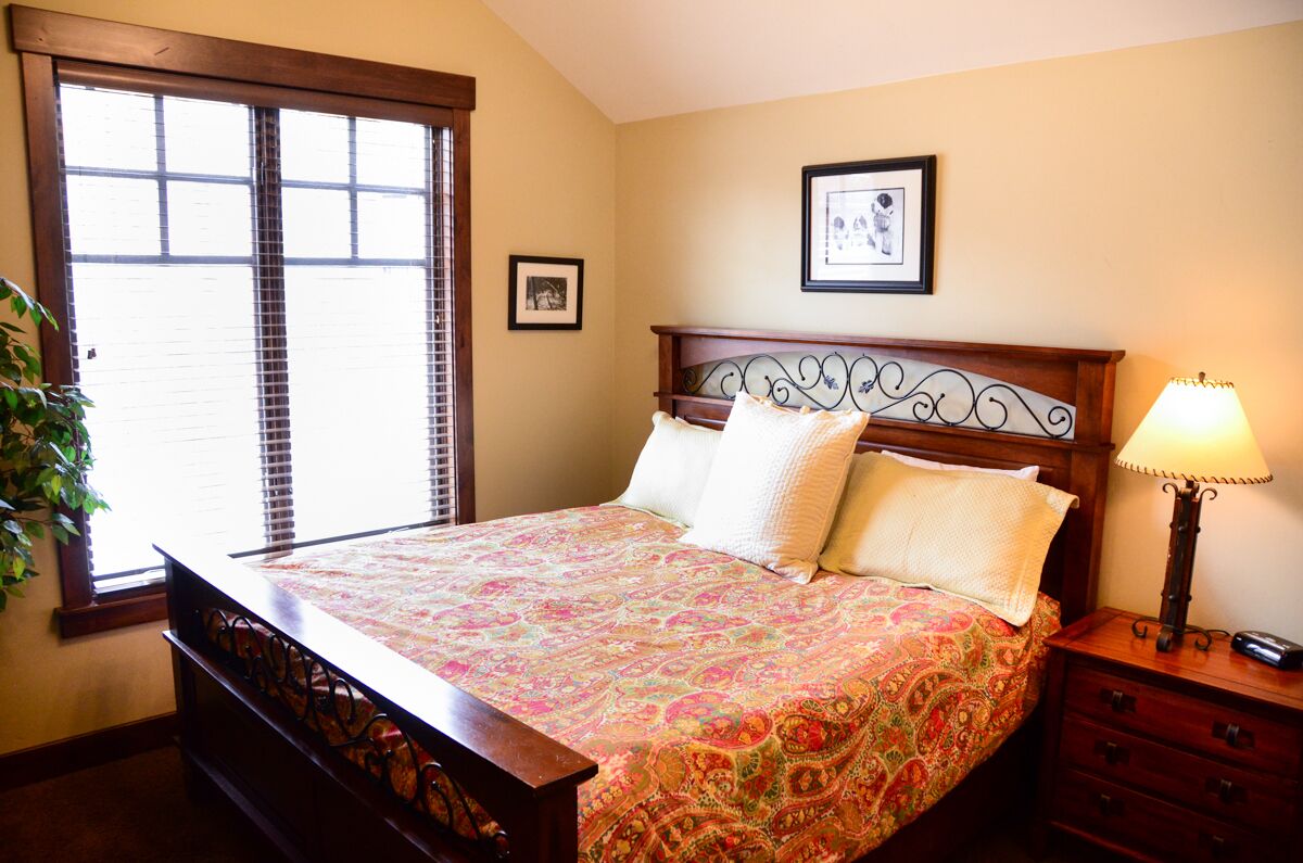 A bedroom with a bed, a nightstand, a lamp, a window with blinds, and a framed picture on the wall.